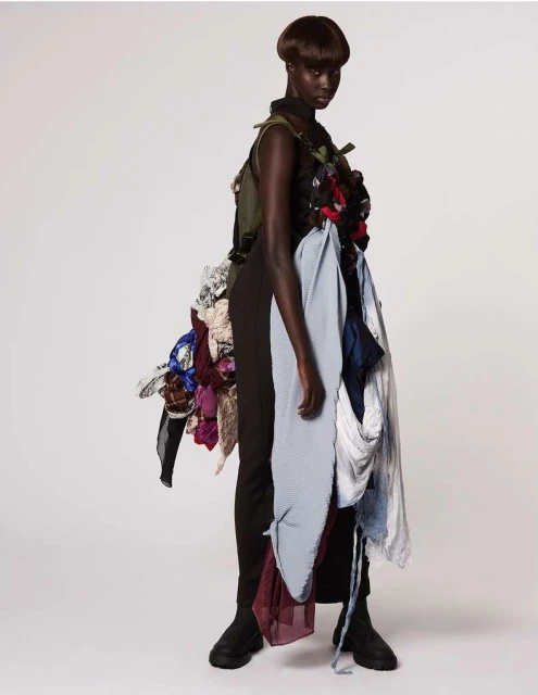 Model showcases avant-garde fashion with layered fabrics and textures, set against a clean white backdrop in a studio setting.