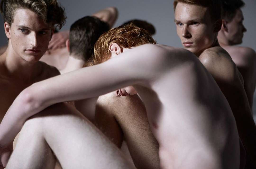 A group of shirtless young adult males huddled together in a close-up composition, creating a sense of intimacy and unity against a light grey background.
