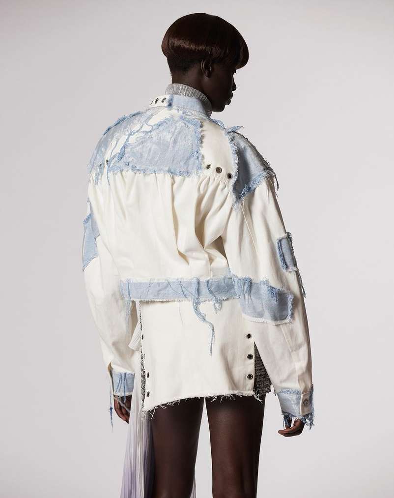 Adult female with dark hair stands in a photo studio wearing a white jacket with light blue patchwork details and a short skirt.