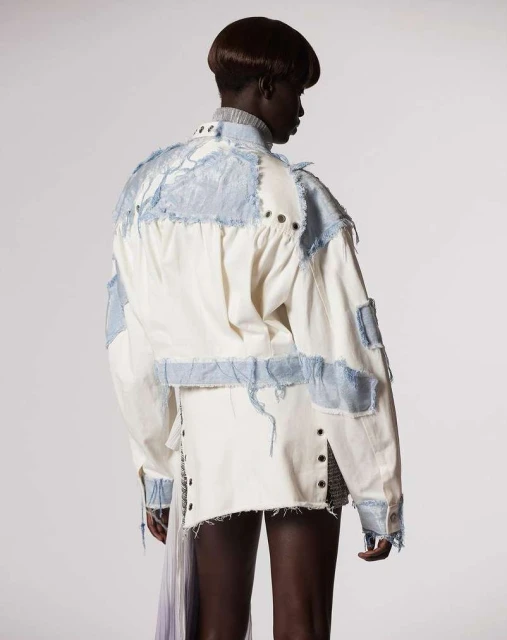 Adult female with dark hair stands in a photo studio wearing a white jacket with light blue patchwork details and a short skirt.