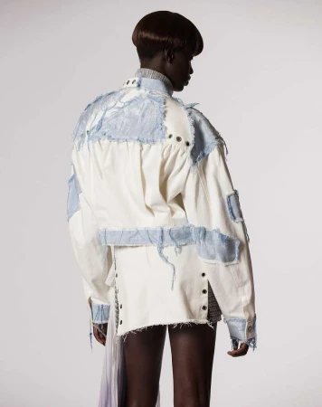 Adult female with dark hair stands in a photo studio wearing a white jacket with light blue patchwork details and a short skirt.