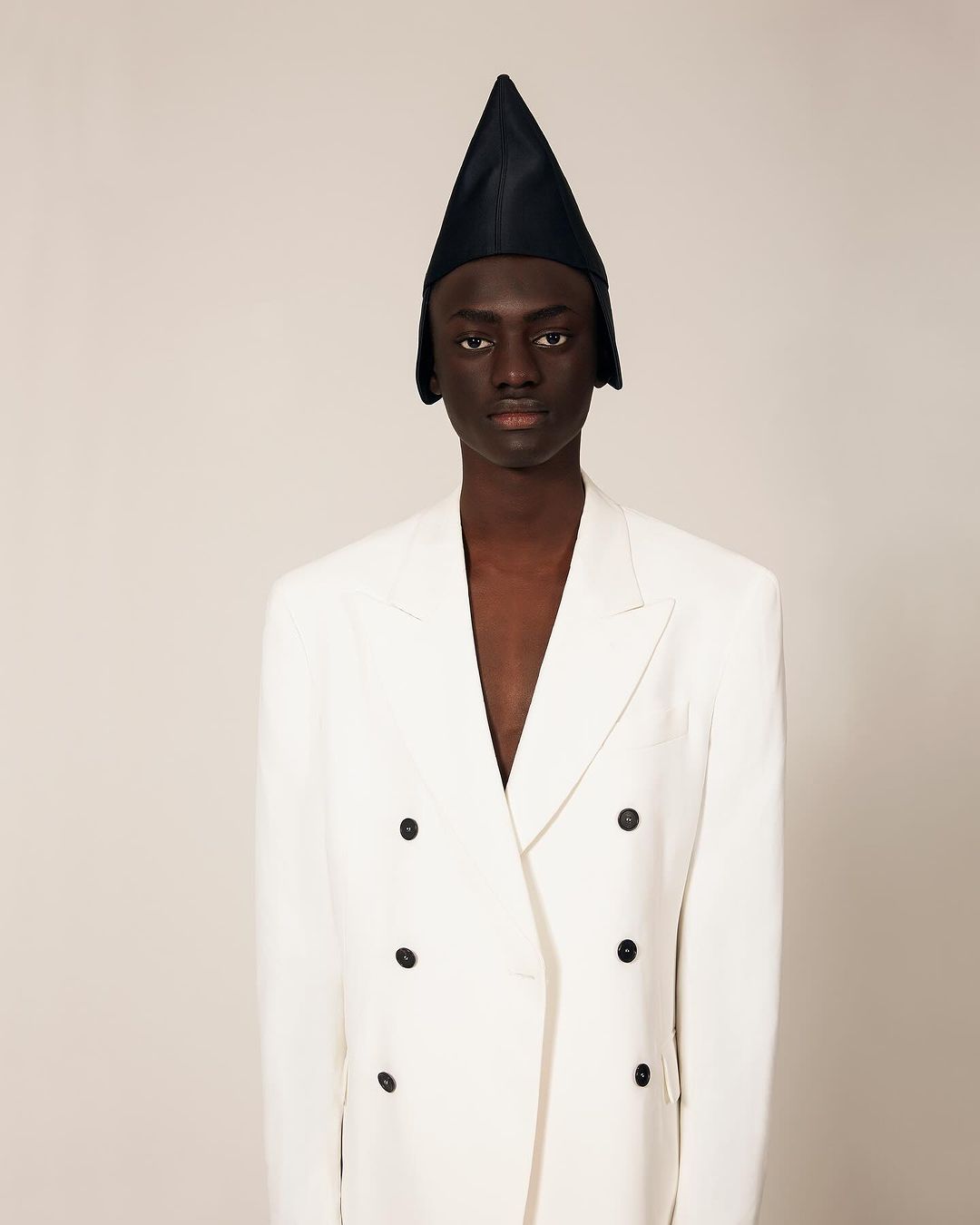An androgynous Black model poses in a photo studio, wearing a white double-breasted blazer and a pointed black hat. The background is neutral.