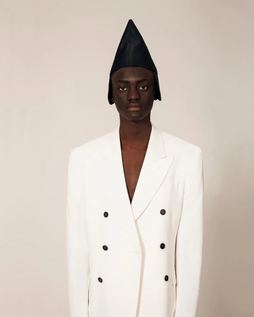 An androgynous Black model poses in a photo studio, wearing a white double-breasted blazer and a pointed black hat. The background is neutral.