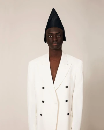 An androgynous Black model poses in a photo studio, wearing a white double-breasted blazer and a pointed black hat. The background is neutral.