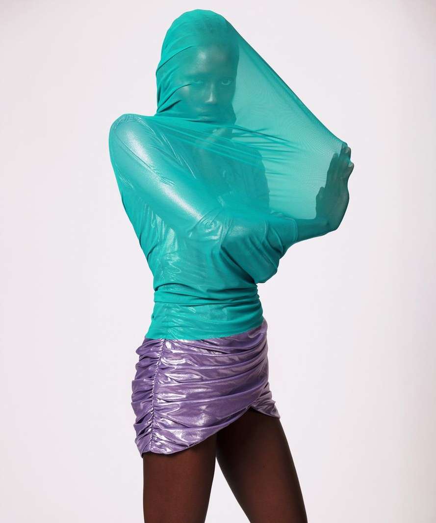 Female fashion model encased in translucent turquoise fabric, wearing a metallic purple skirt against a stark white backdrop in a studio shot.