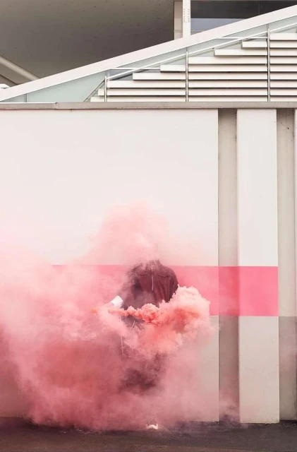 Person stands amidst pink smoke cloud outdoors by a concrete building, creating an atmospheric visual effect, captured in soft light.
