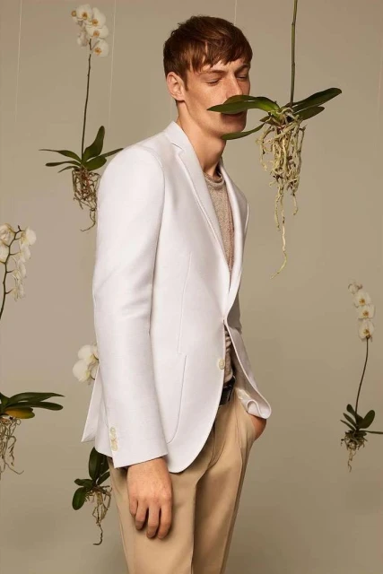 A man in a white suit jacket stands indoors surrounded by orchids, creating a fashionable and elegant scene with a focus on nature and style.