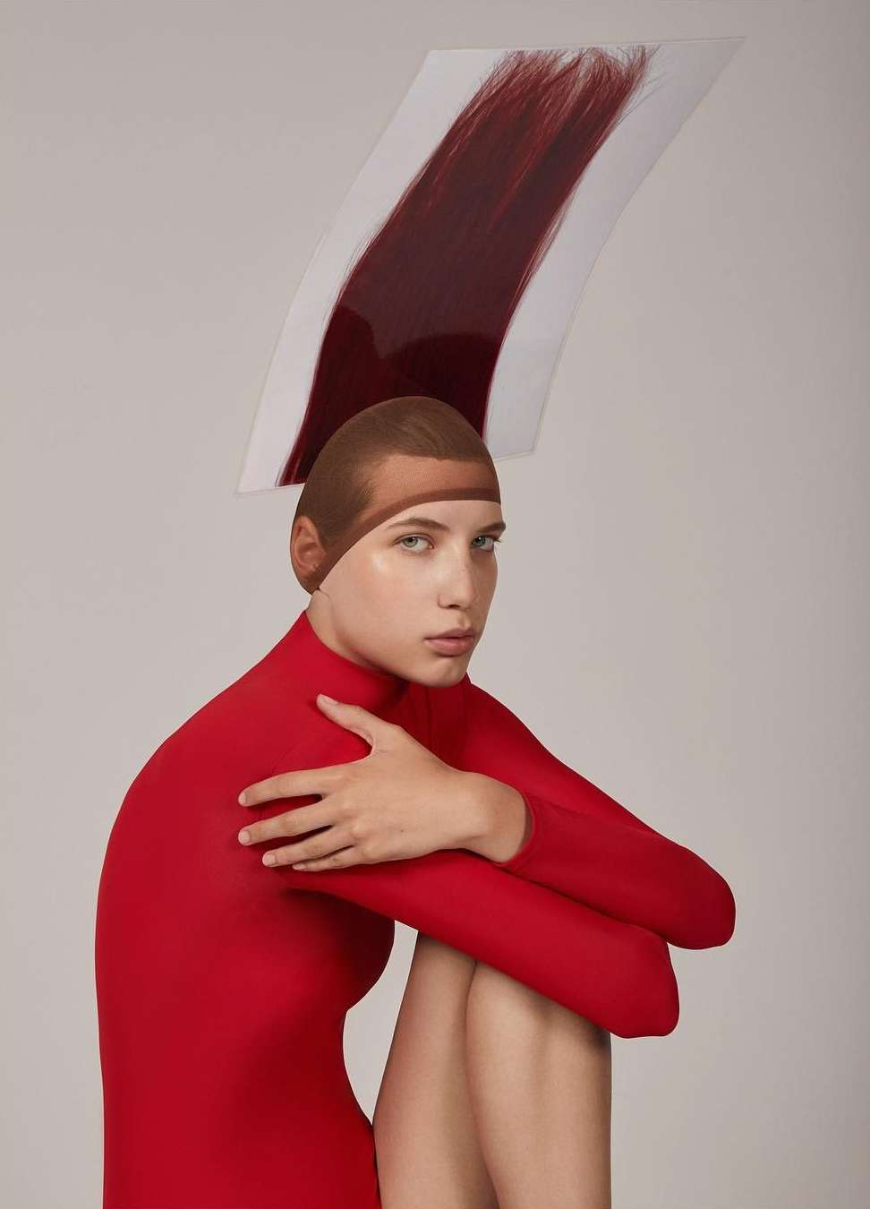 Young woman in a red bodysuit with a tan-colored cap, seated with arms crossed, is seen against a plain backdrop with a swatch of auburn hair above her head.