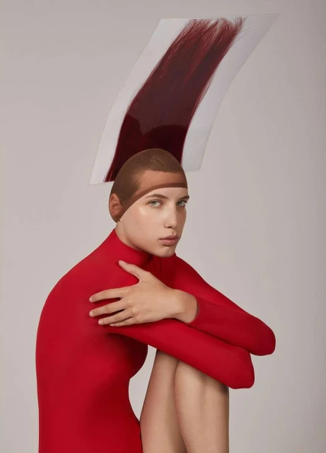 Young woman in a red bodysuit with a tan-colored cap, seated with arms crossed, is seen against a plain backdrop with a swatch of auburn hair above her head.