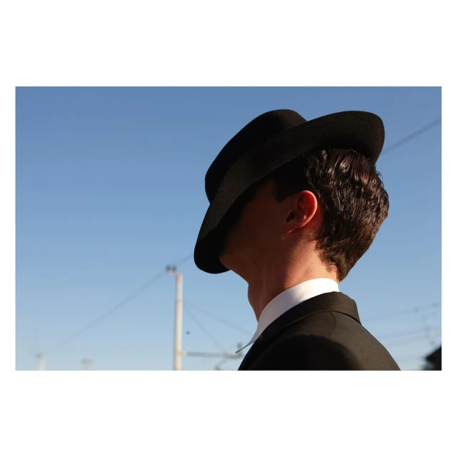 Head and shoulders of a man in a black suit and hat, shown from behind against a clear blue sky, with a power pole in the distance.