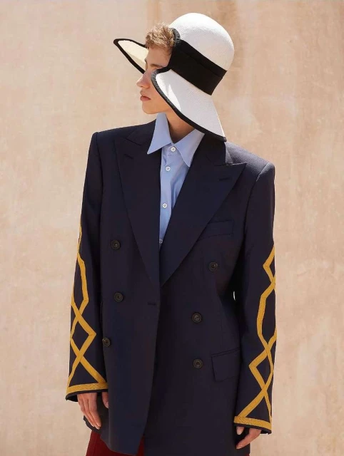 Portrait of a fair-skinned young adult in a wide-brimmed white hat, blue shirt, and oversized navy blazer with gold sleeve accents.