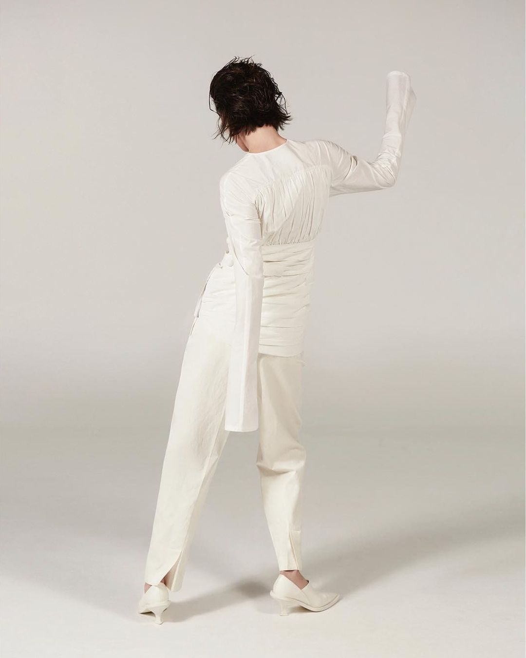 Fashion photo featuring an adult female model posing in a white textured outfit consisting of a long-sleeve shirt, pants, and high heels in a studio.