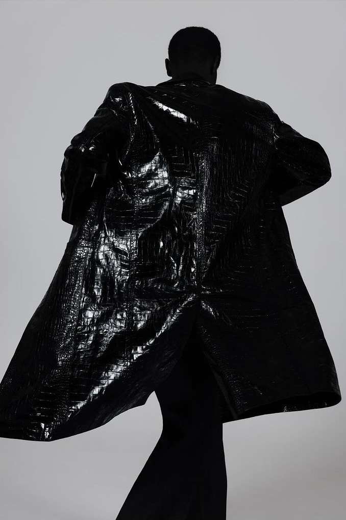 Rear view of a person wearing a shiny, embossed black coat. Fashion editorial shot in front of a plain white background.