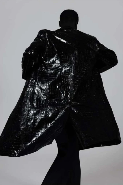 Rear view of a person wearing a shiny, embossed black coat. Fashion editorial shot in front of a plain white background.