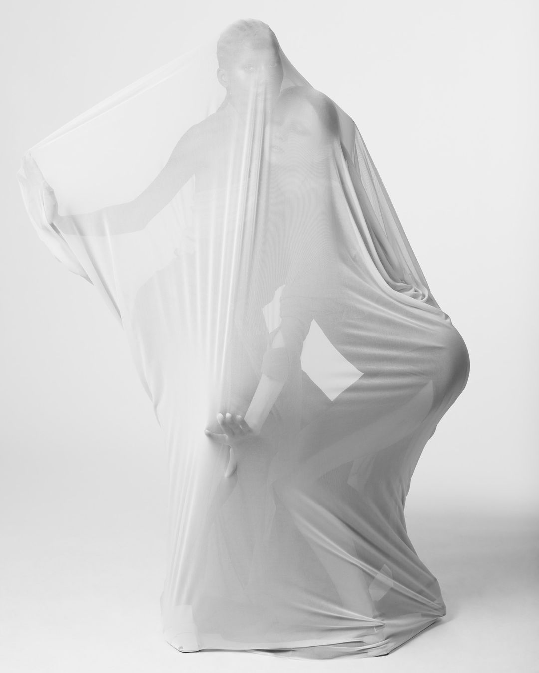 Monochromatic studio shot of a person obscured by a sheer white fabric, creating dynamic shadows and emphasizing abstract form and texture.