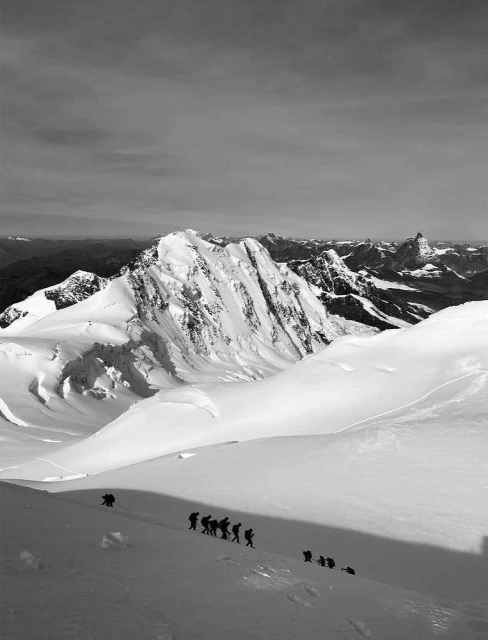 Monochrome landscape shot of a group of hikers ascending a snow-covered mountain, with a view of snow-capped peaks under a cloudy sky.