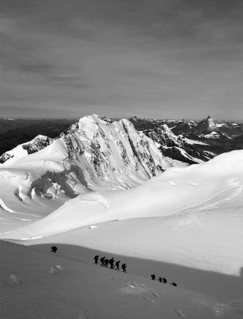 Monochrome landscape shot of a group of hikers ascending a snow-covered mountain, with a view of snow-capped peaks under a cloudy sky.