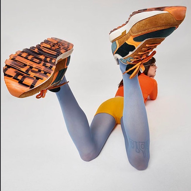 Two athletic shoes are displayed up in the air, with a person wearing colorful leggings and a matching top in the background.