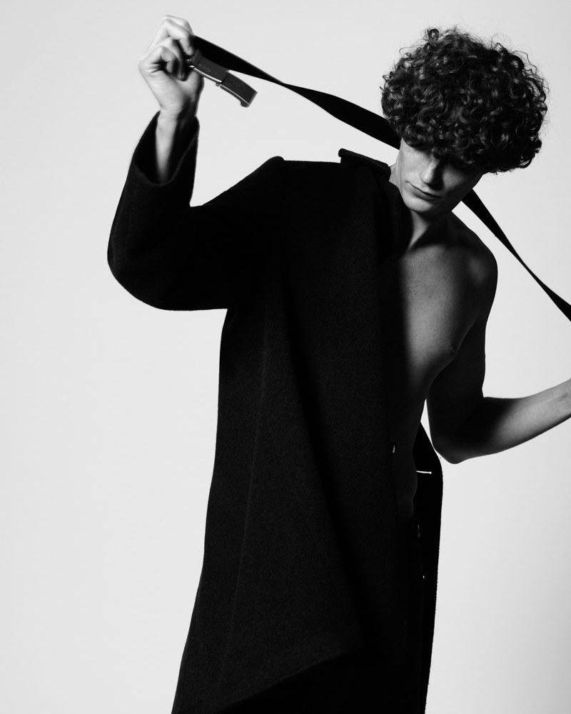 Monochrome portrait of a slender adult male with curly hair, taking off a dark coat and holding dark straps against a light backdrop.
