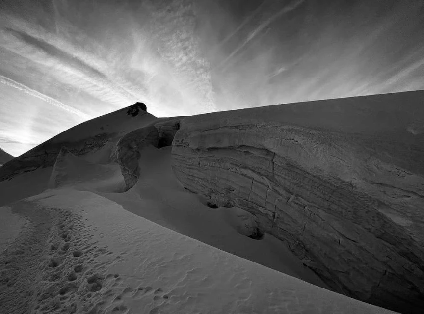 Monochromatic image of snow-covered mountains with striated rock formations, under a sky with high-altitude cirrus clouds and contrails.