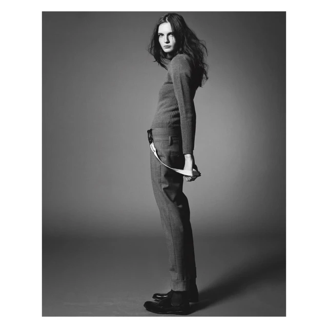 A woman in a grey sweater and trousers stands in a studio with suspenders hanging. Fashion shot with a full length view in black and white.