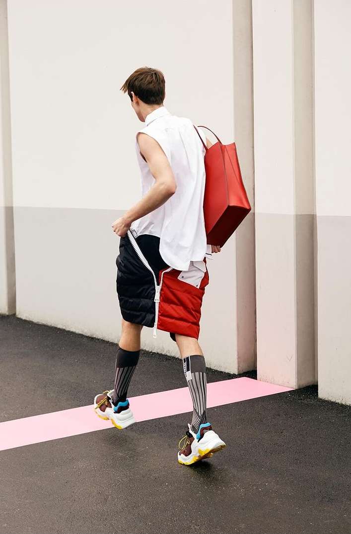 Back view of a stylish adult male walking with a red tote bag, wearing a white top, unique color-block shorts, high socks, and sneakers.