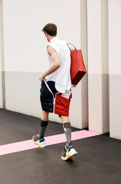 Back view of a stylish adult male walking with a red tote bag, wearing a white top, unique color-block shorts, high socks, and sneakers.