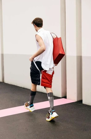 Back view of a stylish adult male walking with a red tote bag, wearing a white top, unique color-block shorts, high socks, and sneakers.