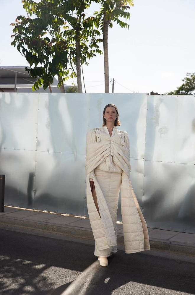 A woman in a puffy, avant-garde off-white outfit poses outdoors against a light-colored wall on a sunny day in a fashion editorial.