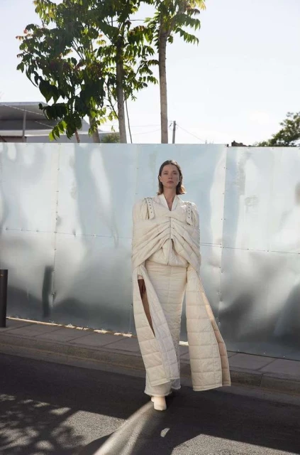 A woman in a puffy, avant-garde off-white outfit poses outdoors against a light-colored wall on a sunny day in a fashion editorial.