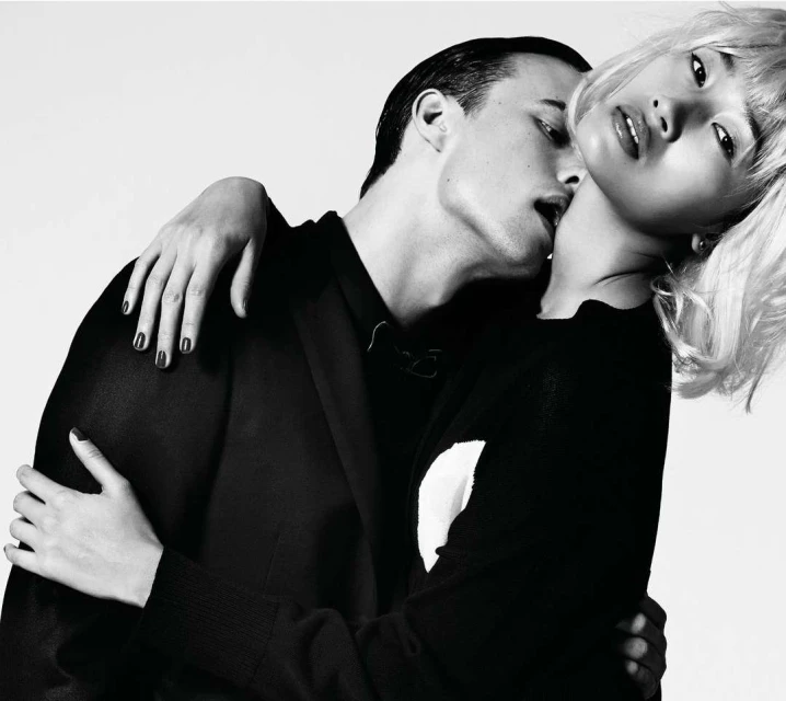 Monochrome fashion shot of a couple. An Asian woman with blond hair leans back as a man embraces her, biting her neck against a white background.