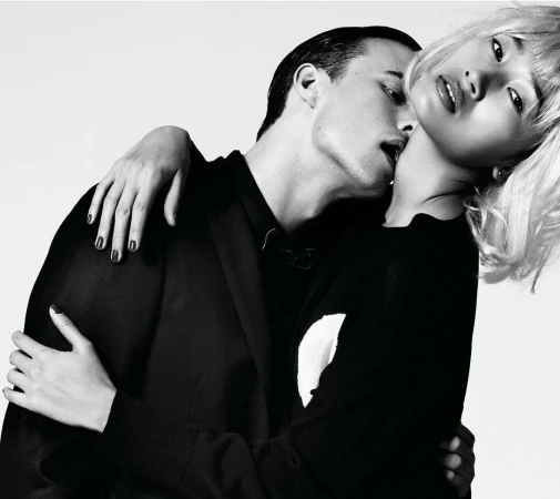 Monochrome fashion shot of a couple. An Asian woman with blond hair leans back as a man embraces her, biting her neck against a white background.