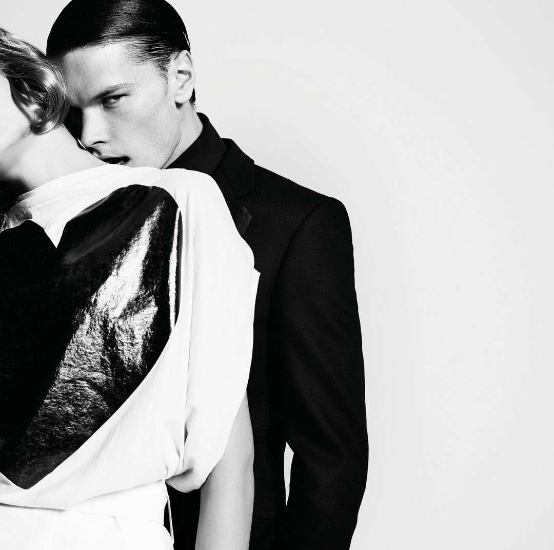A man in a suit is standing behind a woman in a white top with a large black heart in a monochrome fashion shot against a white backdrop.