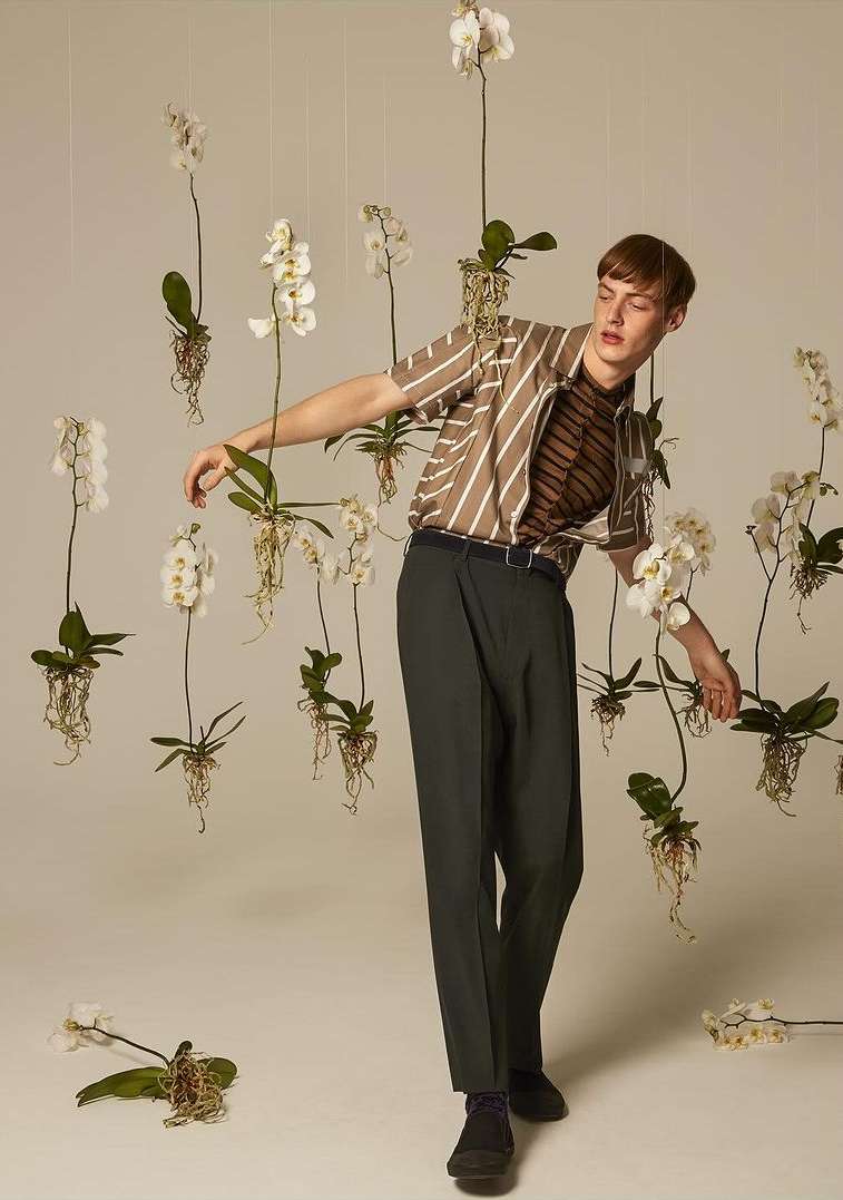 A young adult man poses in front of a backdrop of white orchids while wearing a vertically striped shirt and dark green pants in studio.