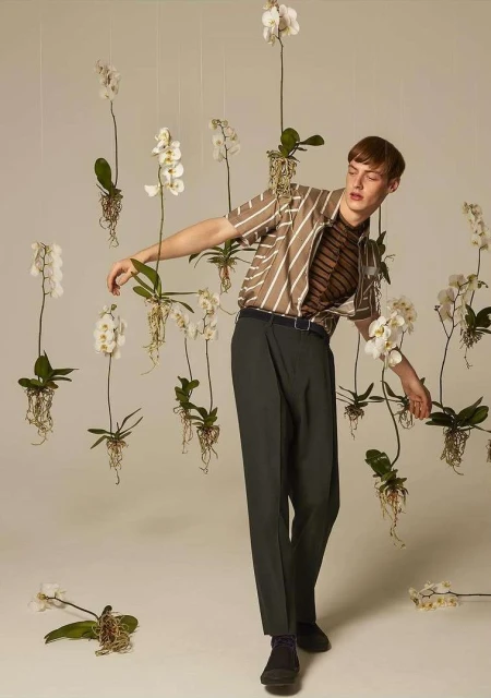 A young adult man poses in front of a backdrop of white orchids while wearing a vertically striped shirt and dark green pants in studio.