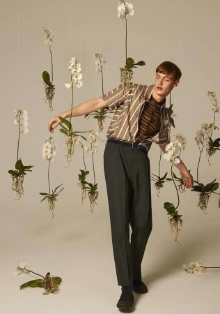 A young adult man poses in front of a backdrop of white orchids while wearing a vertically striped shirt and dark green pants in studio.