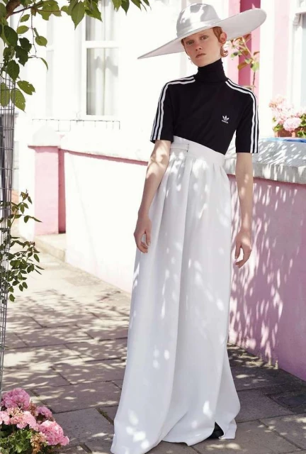 A woman stands in front of a pink-and-white wall, wearing an Adidas top, a white hat, and long white pants. There are pink flowers nearby.