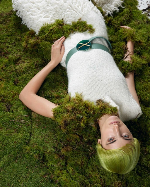 Adult female model in long white dress lying on a bed of moss wearing a green belt and with styled green hair looking at camera.