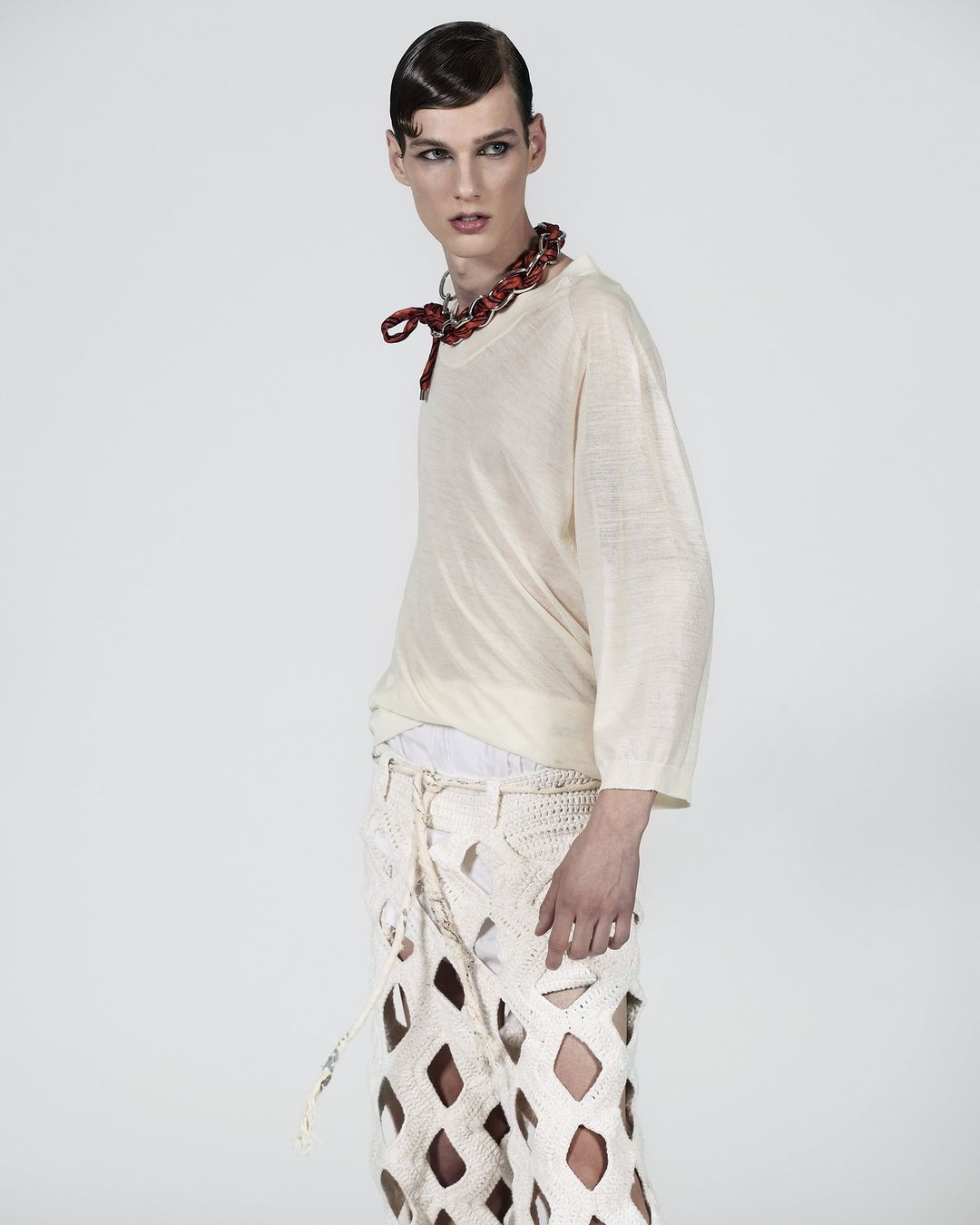 Androgynous male fashion model poses in studio wearing a light-beige top, deconstructed crocheted pants, and a red beaded necklace.