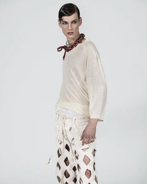 Androgynous male fashion model poses in studio wearing a light-beige top, deconstructed crocheted pants, and a red beaded necklace.