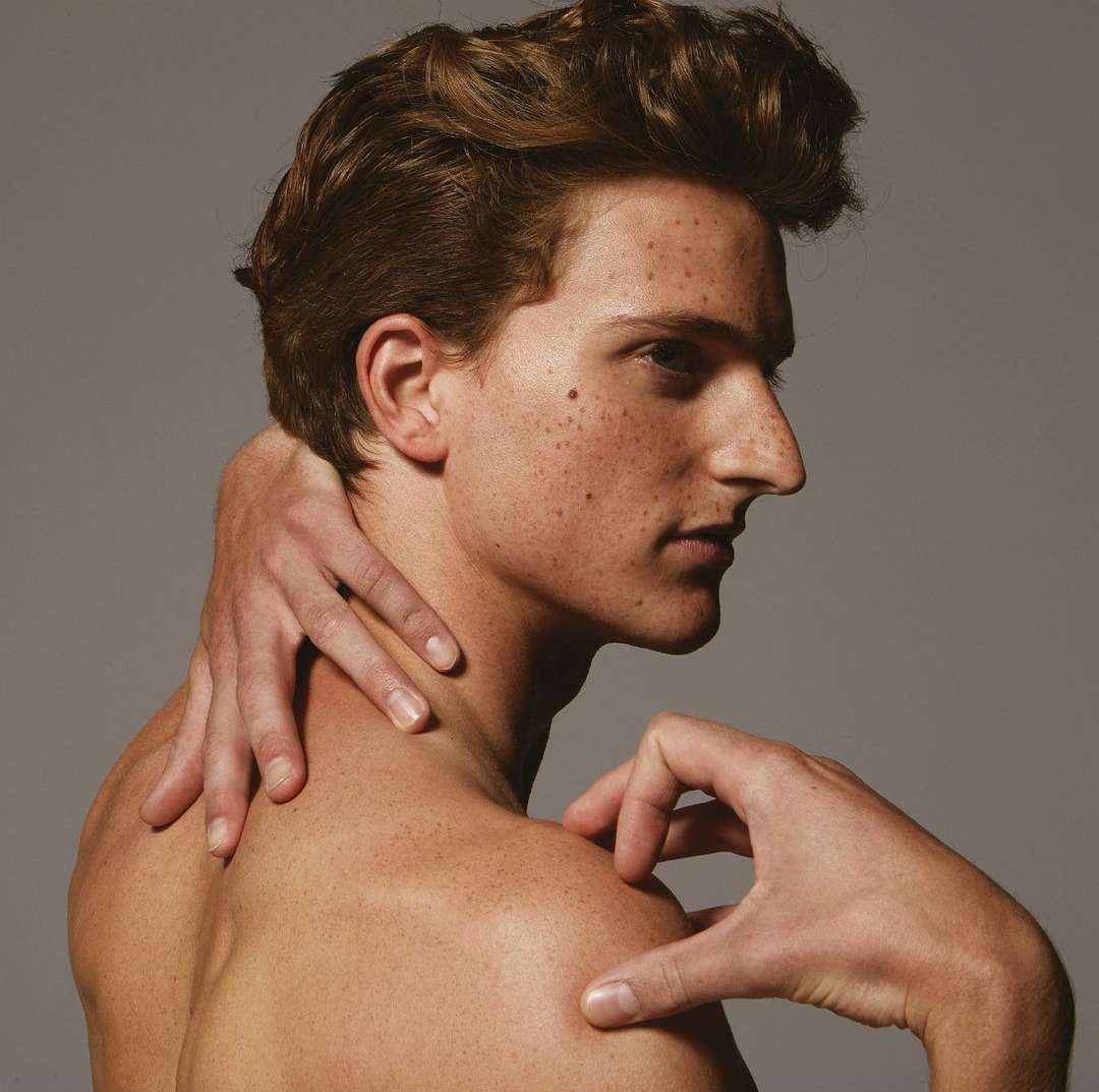 Close-up portrait of a young man with freckles touching his neck and shoulder against a grey background.
