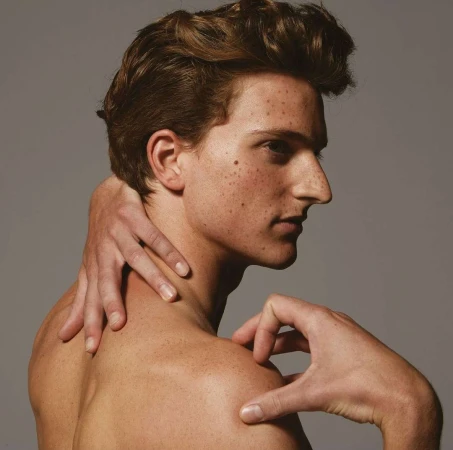Close-up portrait of a young man with freckles touching his neck and shoulder against a grey background.
