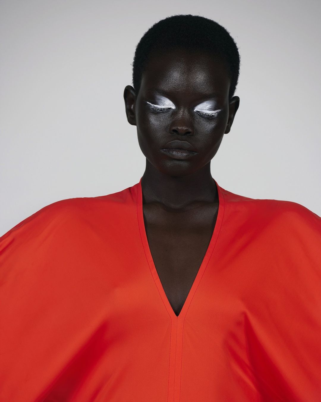 Headshot of a dark-skinned woman with white makeup on her eyelids, wearing a large, loose, vibrant red top against a plain, light backdrop.