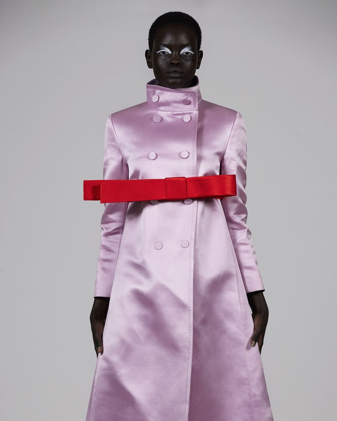 Fashion model wearing a pink coat with a red bow, posing against a light gray background, in a studio shot emphasizing clothing and style.