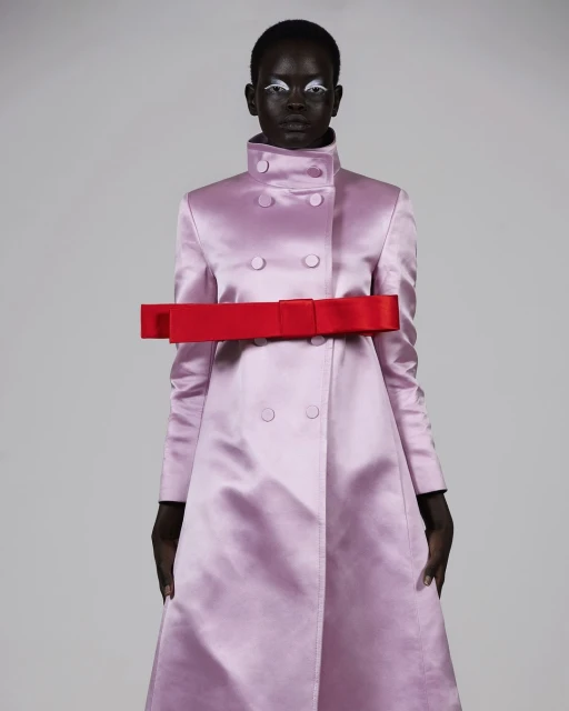 Fashion model wearing a pink coat with a red bow, posing against a light gray background, in a studio shot emphasizing clothing and style.