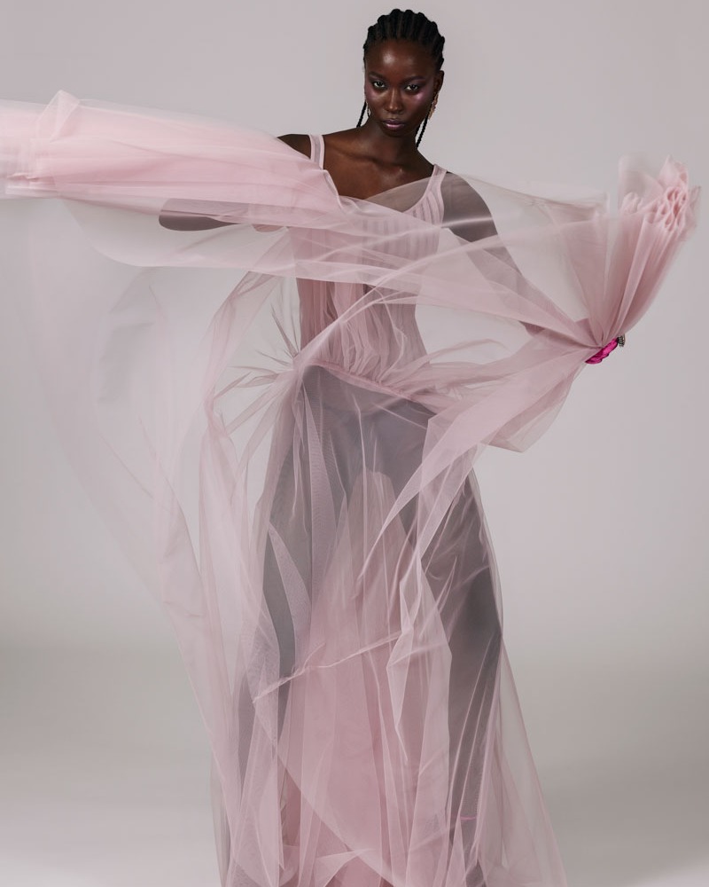 Fashion shot of Black woman in a flowing pink dress and matching scarf against a grey backdrop, captured in a full shot.