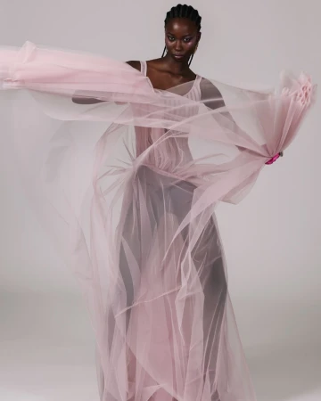Model in a flowing sheer pink tulle dress with arms outstretched against a neutral seamless studio background