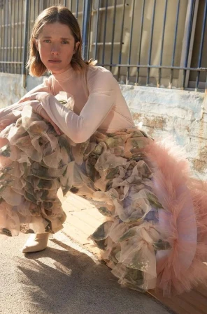 Woman wearing sheer pink top and voluminous ruffled tulle skirt crouching outdoors against a stone wall in Paris.