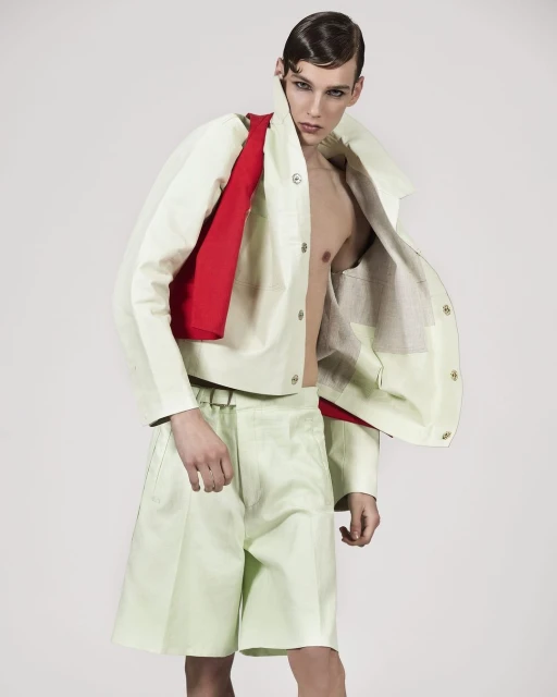 Fashion shot of a young adult male model with a retro hairstyle, wearing a light green jacket and shorts with a red bag.