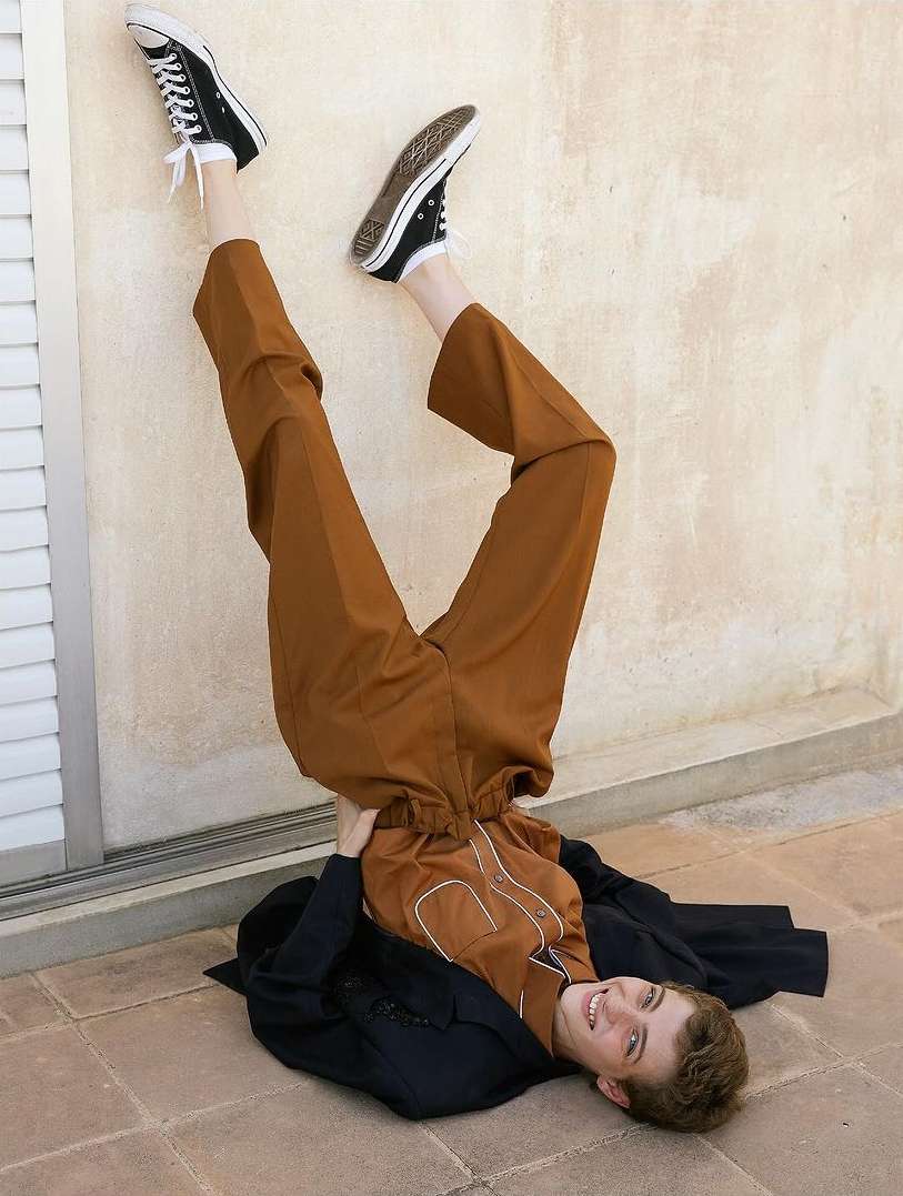 Person in upside down pose wearing brown pants, black sneakers and jacket against off-white wall and tile floor; indoor setting.